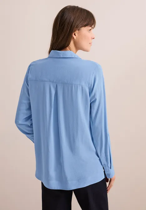 Frau mit hellblauer Bluse und braunen Haaren von hinten, neutraler Hintergrund.
